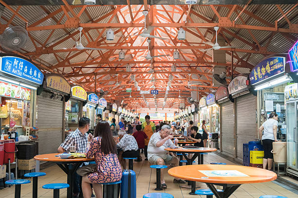 A bustling hawker center in Singapore filled with diners at colorful plastic tables, showcasing an array of food stalls and a vibrant atmosphere.
